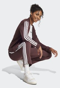 Burgunderfarget treningsdrakt med hvite stripe detaljer, bestående av en jakke med glidelås og tettsittende bukser, kombinert med hvite sneakers. Glatt, lett stoff.