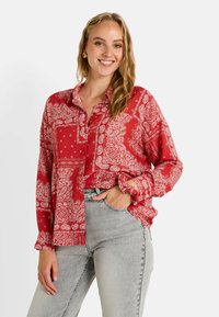 Blouse rouge à manches longues, boutonnée, avec un motif paisley et une coupe loose. Portée avec un jean gris clair, affichant un style décontracté.