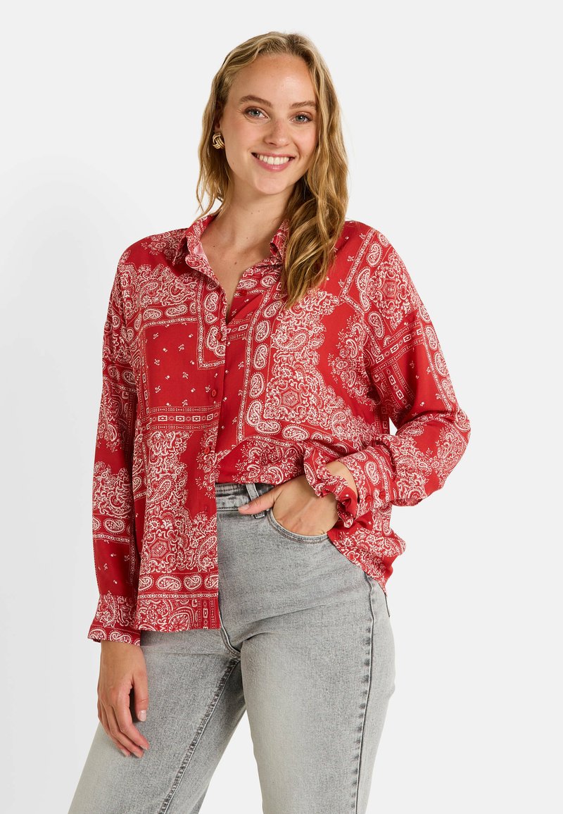 Blouse rouge à manches longues, boutonnée, avec un motif paisley et une coupe loose. Portée avec un jean gris clair, affichant un style décontracté.