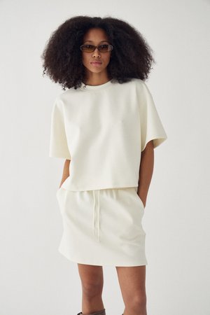 Kvinde med stort krøllet hår iført farvede briller, en løs hvid kortærmet top og matchende hvid nederdel med hænderne i lommerne.