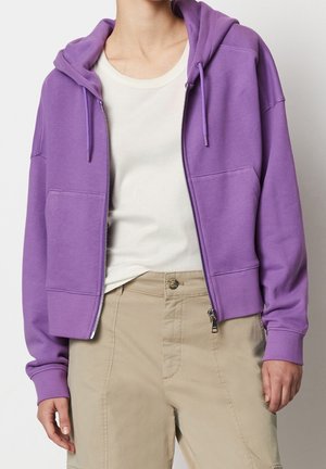 Persona que lleva una sudadera con capucha y cremallera de color lila claro sobre una camiseta blanca y pantalones beige, de pie contra un fondo blanco liso.