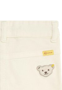 Pantaloni di cotone bianchi con ricamo di un volto d'orso sulla tasca frontale e un'etichetta gialla con testo rosso attaccata alla vita.