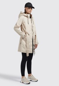 Beige parka takki, jossa on huppu, etuvetoketju, napit ja vyötärösolki. Yhdistetty mustiin legginsseihin ja beigeihin lenkkareihin.