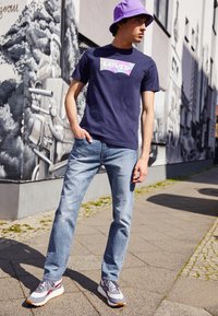 Lila vödörkalap, sötétkék póló színes Levi's grafikával, világoskék farmernadrág és szürke sportcipő piros díszítésekkel, szabadtéri környezetben.