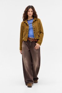 Brūns korduroja žakets ar pogām, zils krekls ar drukātu tekstu, brūni plati bikses un brūnas apavus. Modelis stāv pretī baltai fonam.