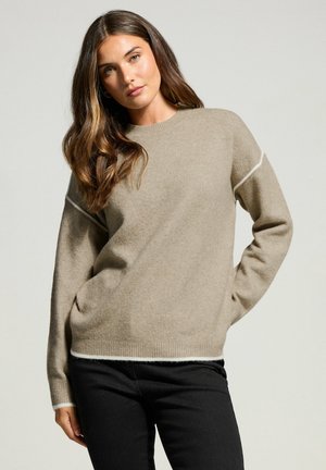 Beige stickad tröja med rund halsringning och långa ärmar, med vita detaljer på muddarna och fållen. Mjuk textur och avslappnad passform.
