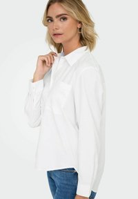 White cotton shirt with a pointed collar, single chest pocket featuring subtle embroidery, and long sleeves, paired with blue jeans.