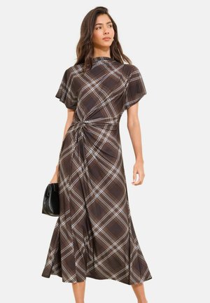 Žena v hnedom károvanom midi šatách s krátkymi rukávmi a uzlovým detailom, držiaca malú čiernu kabelku, pozerajúca sa do strany.