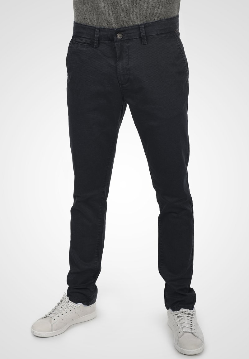 Pantalons noirs en coton au fit slim, avec fermeture à bouton, poches avant et texture lisse, associés à des baskets blanches pour le contraste.