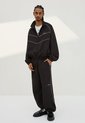 Muž s copánky, oblečený v černé teplákové soupravě s bílým lemováním a černými botami, stojící před světlým jednobarevným pozadím.