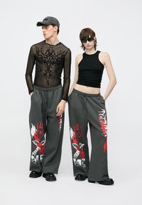 Zwei Models tragen kontrastierende Outfits: ein durchsichtiges schwarzes Langarmshirt mit floralem Muster und ein eng anliegendes schwarzes Tanktop, kombiniert mit grauen Sweatpants mit roten grafischen Designs.