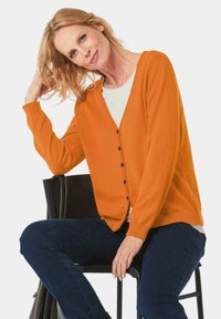 Oranje cardigan met een v-hals, knopen aan de voorkant, geribbelde manchetten en zoom, gedragen over een witte top. Model zit op een zwarte stoel, spijkerbroek zichtbaar.