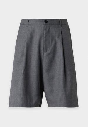 Mand iført en brun T-shirt med lyseblå kant, grå knælange shorts, hvide sokker og sorte sneakers stående med den ene hånd i lommen.
