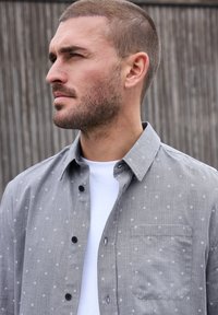Camisa gris con un patrón y cuello abotonado, bolsillo frontal y botones negros, superpuesta sobre una camiseta blanca. Tejido texturado con un diseño sutil.