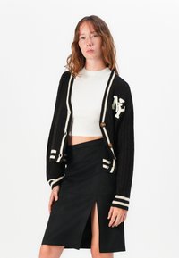 Musta neulecardigan, jossa on valkoiset raidat, napitus ja logo. Yhdistetty mustaan hameeseen, jossa on edessä halkio.