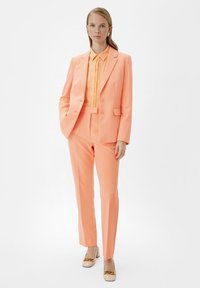 Różowo-brzoskwiniowy garnitur z dopasowaną marynarką i slim-fit spodniami, zestawiony z jasnopomarańczową koszulą i beżowymi butami na słupku.