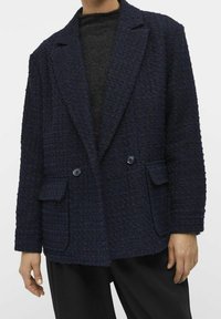 Blazer texturé bleu marine en tissu tissé, présentant un design à double boutonnage, un col à revers et deux poches avant.