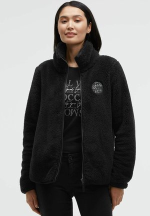 Black fleece jacket with high collar and zip closure. Features a circular logo patch on the left chest. Worn over a black graphic t-shirt.