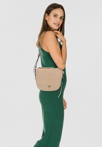 Beige Leder Umhängetasche mit gebogener Form, mit Reißverschlussverschluss und verstellbarem Riemen. Verfügt über dekorative Beschläge und eine weiche Textur.