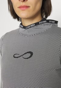 Schwarz-weiß gemustertes Langarmoberteil mit hohem Kragen, das ein schwarzes Unendlichkeitssymbol und einen logogeschmückten Halsausschnitt aufweist.