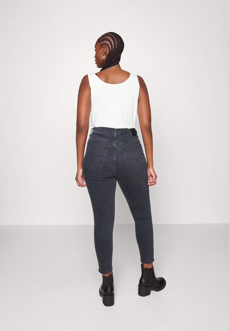Dunkelgraue Skinny-Jeans aus Denim mit hoher Taille, Gesäßtaschen und Gürtelschlaufen, kombiniert mit einem figurbetonten weißen Tanktop und schwarzen Knöchelsockenstiefeln.