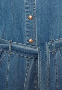 Denimmekko vaaleansinisena, jossa on kuparinapit, vyötärövyö, pystysaumat ja läpätaskut. Kankaan pinta on teksturoitu, mutta sile�ä. 