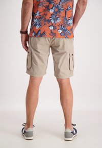 Pantaloni cargo beige con tasche laterali, realizzati in tessuto leggero. Indossati con una camicia a maniche corte con fantasia e sneaker.