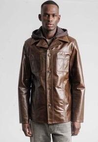 Bruine leren jas met een knoopsluiting, schuin kraag en twee borstzakken. Draag gedragen over een grijze hoodie. Soepele textuur met een slanke pasvorm.