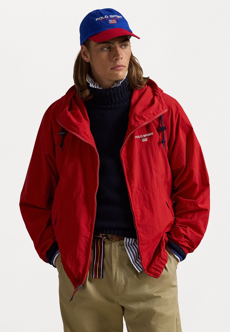 Κόκκινο ελαφρύ μπουφάν με φερμουάρ και κουκούλα, συνδυασμένο με ναυτικό τζέρσεϊ. Η μπλε και κόκκινη καπέλο διαθέτει το λογότυπο 'POLO SPORT'.