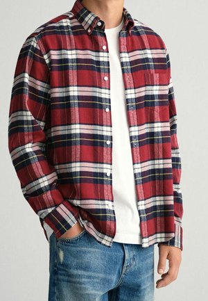 Homme portant une chemise à carreaux rouge, blanche, bleu marine et jaune boutonnée sur un t-shirt blanc avec un jean bleu, main dans la poche.