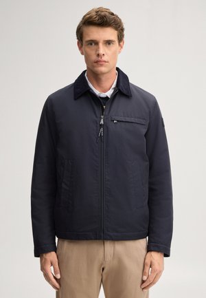 Mann trägt eine marineblaue Jacke mit Reißverschluss und Kragen über einem weißen Hemd und beigen Hosen, steht vor einem schlichten hellen Hintergrund.