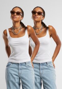 Débardeur blanc à côtes avec des bretelles épaisses, associé à un jean loose bleu clair. Modèle portant des colliers en chaîne argentée empilés et des lunettes de soleil brunes.