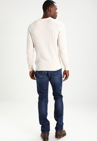 Suéter de punto trenzado crema con puños y dobladillo acanalados, combinado con jeans azul oscuro y zapatos marrones, visto desde la parte posterior.