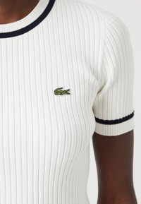 Bílý žebrovaný top s krátkými rukávy, s námořnicky modrým límečkem a manžetami a malým zeleným krokodýlím logem na levé straně hrudi.