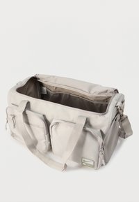 Ανοιχτό μπεζ τσάντα duffel με διπλές λαβές, δύο μπροστινές τσέπες με φερμουάρ, μια πλαϊνή τσέπη από δίχτυ, και ένα ανοιχτό επάνω τμήμα σε λευκό φόντο.