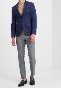 Man som bär en marinblå blazer, beigemelerad tröja, grå slim-fit byxor och svarta läderloafers står mot en vit bakgrund.
