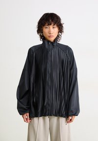 Black pleated jacket with a high collar, zip front, and long sleeves. Features metallic accents along shoulder seams; smooth, glossy texture.