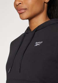 Crna kapuljača od mekanog materijala s kengur džepom, s bijelim Reebok logotipom i špagicama. Jednostavan dizajn s ležernim krojem.