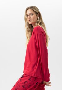 Vrouw met lang blond haar, gekleed in een rode shirt met lange mouwen en rode broek met zwarte cartoonfigurenprints op een witte achtergrond.