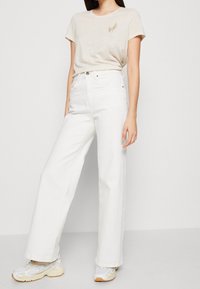 Camiseta de algodón beige con un pequeño bordado de hoja, combinada con jeans de pierna ancha blancos de cintura alta y zapatillas grises con un diseño sutil.