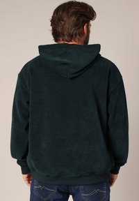 Hoodie en velours côtelé vert foncé avec une grande capuche, texture cannelée et poignets élastiques, associé à un jean bleu avec des coutures sur les poches arrière.