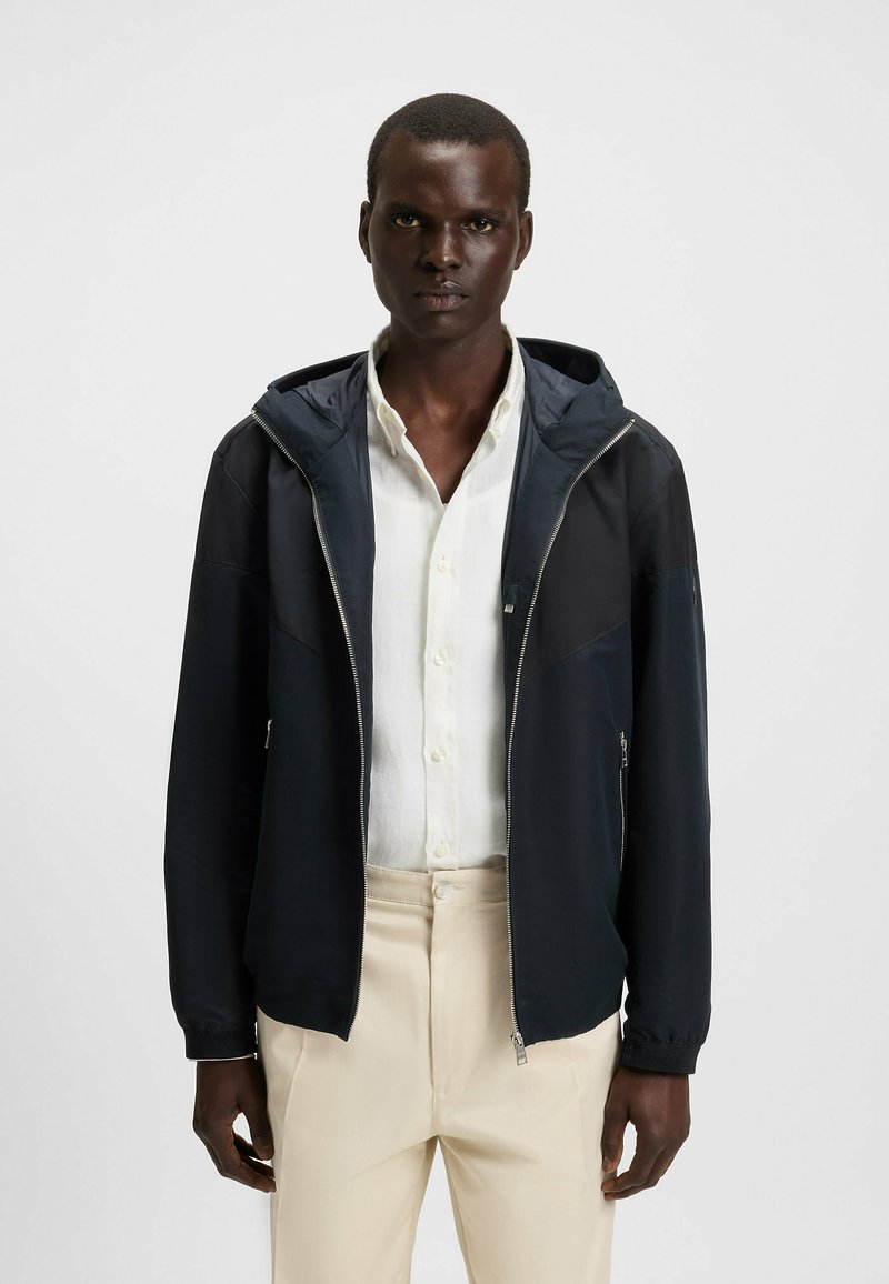 Chaqueta con capucha de color azul marino y cierre frontal, con detalles en panel, puños elásticos y una textura suave, llevada sobre una camisa blanca y pantalones beiges.