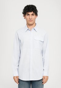 Mann med krøllete mørkt hår iført en lyseblåstripet langermet skjorte med knapper og blå jeans, som står mot en ensfarget bakgrunn.