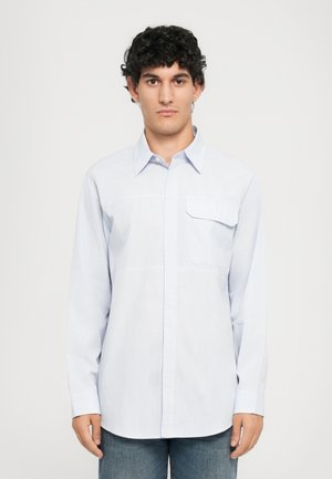 Mann med krøllete mørkt hår iført en lyseblåstripet langermet skjorte med knapper og blå jeans, som står mot en ensfarget bakgrunn.