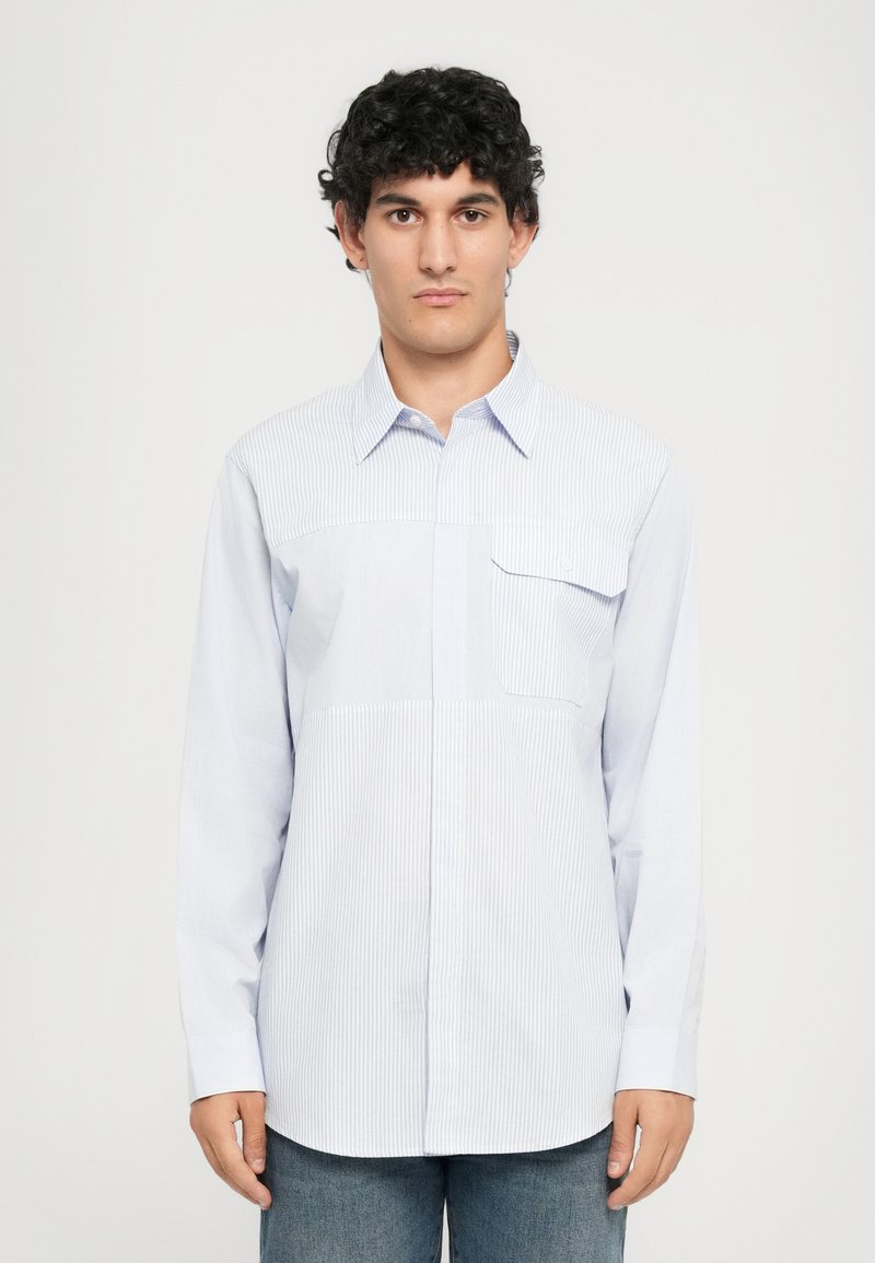Mann med krøllete mørkt hår iført en lyseblåstripet langermet skjorte med knapper og blå jeans, som står mot en ensfarget bakgrunn.