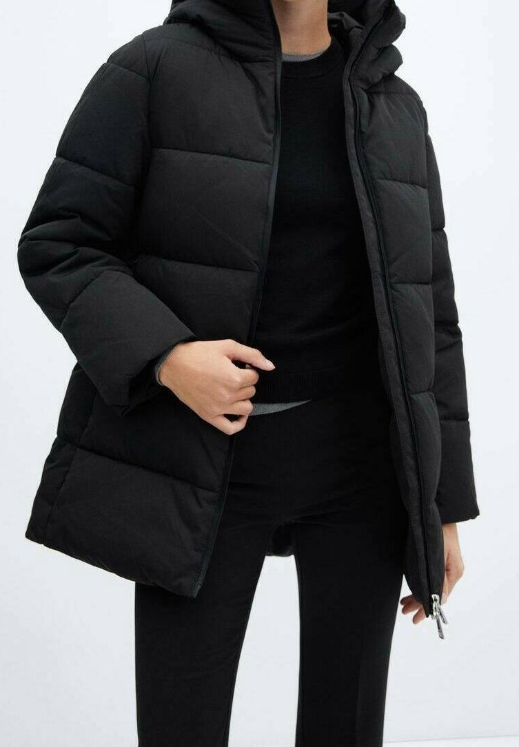 Personne portant une doudoune noire matelassée avec une capuche, un pantalon noir et un haut noir, tenant la fermeture éclair de la veste près de la taille.
