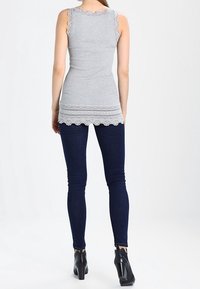 Szary top bez rękawów z falistymi krawędziami, teksturowaną tkaniną i koronkowymi zdobieniami u dołu, zestawiony z ciemnoniebieskimi wąskimi dżinsami i czarnymi botkami za kostkę.