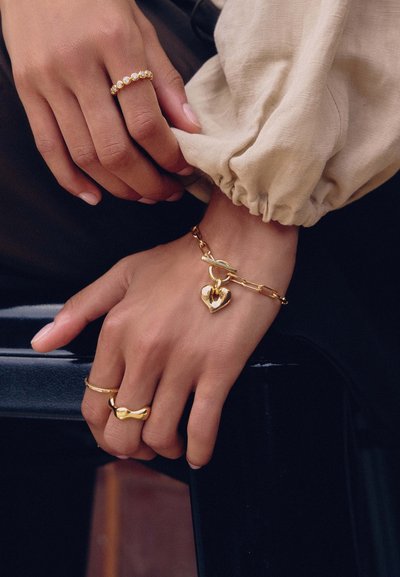 Χέρια με τρία χρυσά δαχτυλίδια και χρυσό βραχιόλι αλυσίδα με κρεμαστό καρδιάς, ακουμπισμένα σε μαύρη επιφάνεια, μερικώς ορατό μπεζ μανίκι.