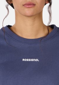 Niebieska bluza z prążkowanym dekoltem, z białym logo "ROSSIGNOL" na przodzie. Materiał wydaje się być miękki i teksturowany.