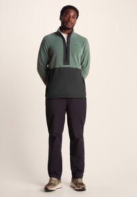 Green fleece pullover with a black lower half, half-zip front, and stand collar. Paired with dark pants and light sneakers.